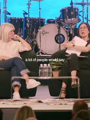 A low-angle shot captures two women seated on a platform, engaged in conversation. Behind them, a drum set and sound equipment are visible, suggesting a stage or performance setting. Text overlaid states, 'a lot of people would say'. The composition includes a table with water bottles, enhancing the context of a formal discussion.