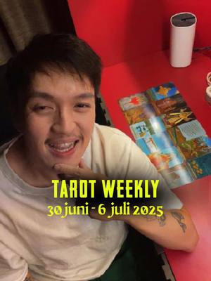 A man with short dark hair and a white t-shirt smiles, resting his chin on his hand. The background is red with a white cylindrical object on a table. On the table is the tarot cards that has been opened. A text overlay in yellow font says "TAROT WEEKLY" and "30 juni - 6 juli 2025".