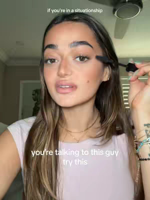 A woman with long, dark hair and makeup is shown in a close-up shot, holding a mascara wand near her eyelashes. She appears to be applying makeup, and the background is softly blurred, with a ceiling fan visible.