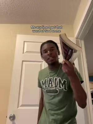 A young Black man in a green t-shirt holds a Bible in front of a white door. The man is in a room lit with a warm yellow color. Text overlayed on the image reads, with a caption that says 'Me anytime I got that word of God on me'.