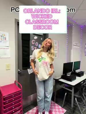 A woman stands in a classroom decorated with decor from 'Wicked: For Good.' The background features an open doorway, a desk with a computer, and a storage cart. Text overlay reads 'Orlando IRL: Wicked Classroom Decor'. The woman wears a graphic tee, jeans, and sneakers, while standing on a pink checkered rug.