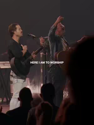 The image is a vertical, dimly lit shot of a live performance or worship service. Two men are on stage, positioned left and center. The man on the left plays an acoustic guitar and wears a dark long-sleeved shirt and light-colored pants. The man on the right, slightly older, has his right arm raised. The crowd is blurred at the bottom. In the center of the image, there is the overlay text 'HERE I AM TO WORSHIP'.
