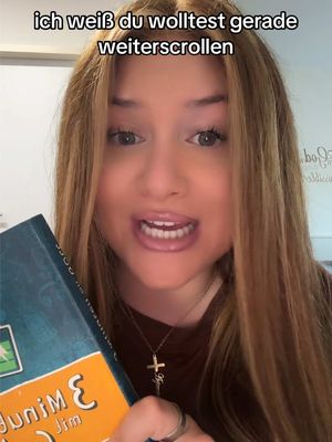 A young woman with long blonde hair, holding a book and speaking directly to the camera. The background is blurred, suggesting an indoor setting. She wears a maroon top and a cross necklace.