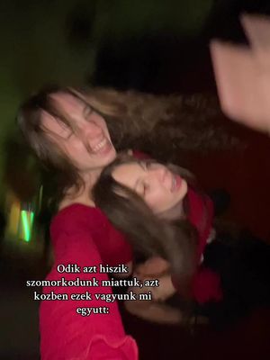 Two young women are embracing and laughing. The woman in front is wearing a red top and smiling widely, with her hair down. The other woman is partially obscured behind her, also laughing. The background is dark, with some out-of-focus visual elements suggesting a crowded indoor setting. The image is slightly blurred, conveying a sense of movement or a candid moment.