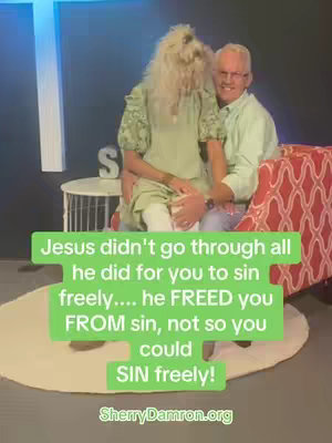 An exterior shot features two figures standing together, likely in a studio setting. A woman in a flowing green dress with long blonde hair sits in the arms of an older man, dressed in a light green shirt and glasses, who is seated on a red patterned couch. A green-colored rectangle overlays the image with the text: "Jesus didn't go through all he did for you to sin freely.... he FREED you FROM sin, not so you could SIN freely!". The background has a blue wall and a white cross.