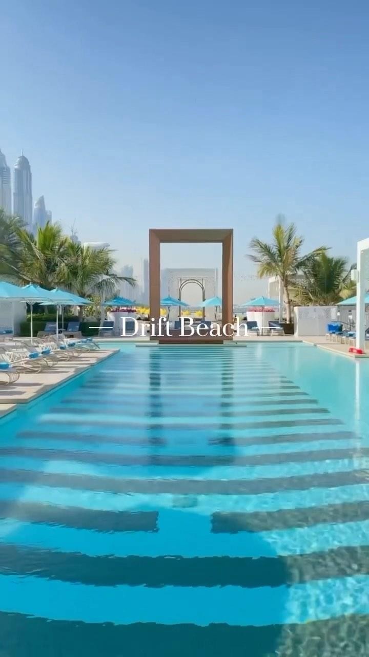 The image shows a serene outdoor scene with a swimming pool as the central focus. The pool has clear blue water and is surrounded by a stone deck with lounge chairs and blue umbrellas. Palm trees and other greenery are visible, as well as a cityscape in the background. Above the pool, there is a large rectangular wooden structure. The sky is clear blue.