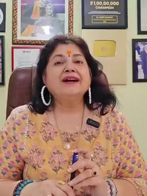 A woman with dark hair and fair skin is centered in the frame, wearing a yellow patterned top and large hoop earrings. She has a red bindi on her forehead. Behind her is a room with framed awards and pictures on the wall. She is looking up towards the camera. The overall composition is well-lit, and the background suggests a professional setting.