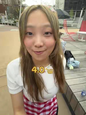 A young woman is captured in a selfie-style shot, appearing outdoors. She has long, light brown hair that is parted on the side. Behind her, the setting includes blurred outdoor elements like trees, a metal deck, and some seating. Her expression is friendly and engaging. Overlayed on the image is the text "4.0" beside a cupcake icon, likely indicating her GPA.