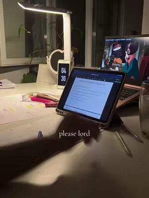 A well-lit desk scene at night. An iPad with a document on it is positioned in the foreground. Papers, pens, and a laptop are laid out. A bedside lamp and a phone are lit up, with the time 04:30 on the phone screen.