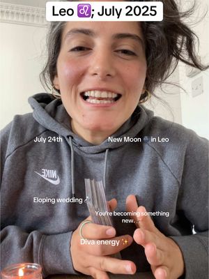 A selfie-style video cover image featuring a woman with graying hair and a gray hoodie in a well-lit setting. She is smiling and holding a deck of cards, with overlaid text indicating astrological reading details. Background elements include interior walls and a glimpse of a lit candle.