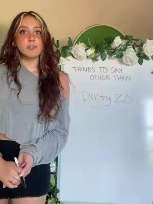 A woman stands in front of a whiteboard, holding a marker. The board has text written on it, a floral arrangement sits behind her, and a green arched element frames the top of the whiteboard.