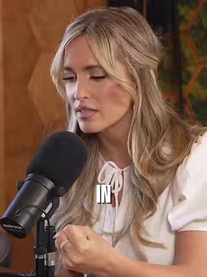 A woman with blonde hair is positioned slightly off-center, facing towards the left and looking down. She is wearing a white blouse with a bow detail. A microphone is in front of her, angled towards her mouth. The background appears to be a wooden wall. A text overlay with the word "IN" is partially visible. The overall composition focuses on the woman, with a soft lighting and neutral background.