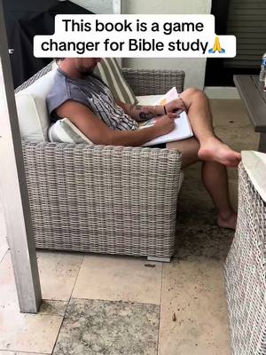 A man sits in a wicker chair outdoors, holding a notebook and pen, with a Bible in front of him. Text overlay at the top reads, "This book is a game changer for Bible study." The background includes the corner of a building and outdoor furniture.