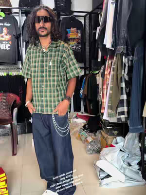 A full body shot of a man in a clothing store. He wears sunglasses and a plaid shirt, holding his hands in his pockets. Jeans and accessories are shown in the background. The floor is tan and the store interior is neatly organized.