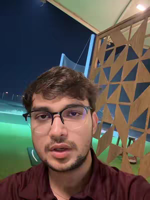 The image shows a medium shot of a man wearing glasses, looking directly at the camera. He has short brown hair and a beard. He is wearing a maroon shirt. The background behind him is out of focus but appears to be an outdoor setting, possibly a golf course, with a decorative wooden lattice structure to the right.