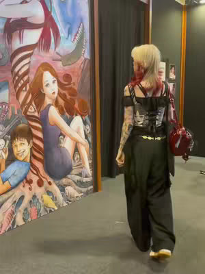 A woman with blonde hair and red highlights is walking away from the camera. She is wearing a black outfit with tattoos visible on her arms. Behind her is a large painting with manga-style art, featuring figures and elements of a Junji Ito artwork. The environment appears to be an exhibition space or convention hall.