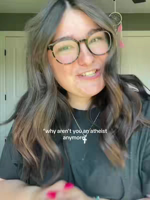 A woman with long brown hair and glasses is smiling at the camera. The background is blurred, but suggests an interior setting with a door and view of outdoor foliage. She is wearing a black top and a necklace with a cross pendant.