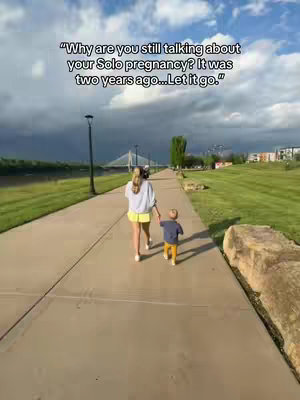 A woman and a young child walk along a paved path, holding hands. The path has green grassy areas on either side. A cloudy sky is overhead with a bridge in the background.