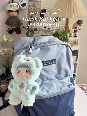 A light-blue Hawk backpack is the central item, positioned against a background of a white surface and various decorative items. A small, plush doll in a mint-green outfit with a bear-like hood is attached to the backpack. The backpack has a Hawk logo visible, and above it the text 'Pack with me' and 'Hawk Backpack Blue-dust/Limoges' text. The setting appears to be a well-lit, organized space, possibly a desk or shelf. The color palette is soft and pastel, dominated by blues, greens, and whites.