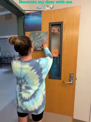 A person is shown from the back decorating a wooden door with various images and cutouts. The person is wearing a tie-dye long-sleeved shirt and black shorts, their hair is pulled up in a bun. The background has a school hallway setting.