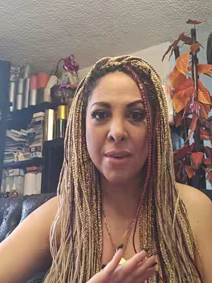The cover image shows a medium shot of a woman with long, braided hair, looking towards the camera. She wears a necklace and has a neutral expression. Behind her, there is a shelf with various items, and a leafy plant adding depth to the background.