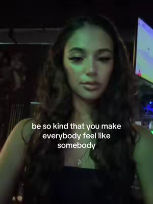 A medium shot shows a young woman looking towards the camera. She has long wavy brown hair and is wearing a black top. Text overlays on the bottom, surrounded by softly blurred interior lights.