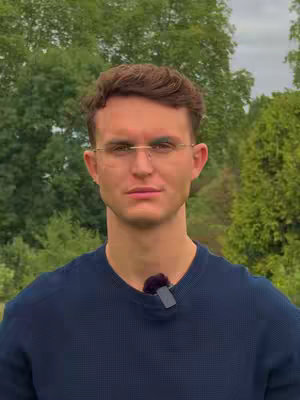 A medium shot of a man wearing glasses and a dark blue sweater, with a lush green outdoor background. He is looking directly at the camera, and a small microphone is clipped to his shirt.