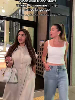 Two women are walking side by side out of a building. The woman on the left is wearing a light beige dress and carrying a white purse. The woman on the right is wearing a white tank top and light blue jeans. The background is a building with glass doors.
