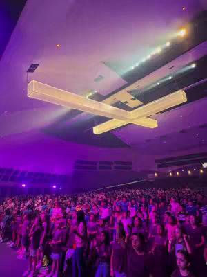 An image of a large, open auditorium filled with people. The ceiling is high, with large, bright lights in a cross shape. The walls are a mix of purples and pinks, suggesting the event is lit for atmosphere or performance. The crowd is densely packed, suggesting a concert, conference, or similar event. People are looking towards a stage or focal point, and their postures vary, indicating active participation and engagement in the event. It's a wide shot that captures the scale of the gathering and the energy of the attendees..