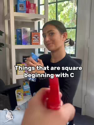 A woman looks at the camera while holding a blue spray bottle. The foreground features a red spray bottle, suggesting an interactive scenario. Bookshelves filled with colorful boxes and writing are in the background.