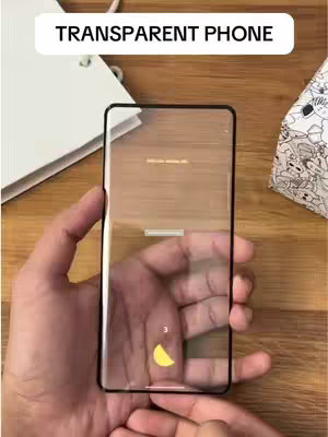 A person's hands holding a transparent smartphone. The phone dominates the frame, displaying what appears to be a loading screen with a yellow semi-circle. The background is a light brown wooden surface, with a white notebook and a patterned box in the top corners adding visual interest. The overall composition focuses on the tech gadget against a simple setting.