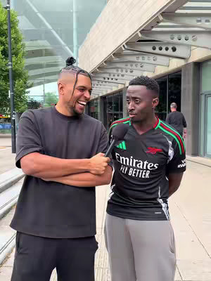 Two young men stand outside against a backdrop of a modern building. The man on the left, with a high bun, smiles broadly while holding a microphone, positioned as if he is interviewing the man on the right. This man has an Arsenal football shirt on and looks at the microphone. They are standing on paving stones in front of glass doors by a building.
