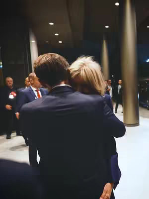 An image shows one person embracing another from behind. The person in front is wearing a dark blue suit with the back view visible. The person being embraced has blonde hair and their face is partially hidden. They appear to stand in a hall with several people in the background, some in suits. The lighting is dim, creating a sense of privacy.