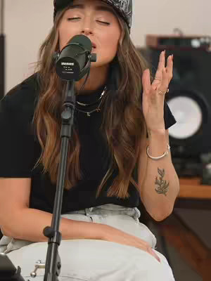 A woman with long brown hair and a black cap sings into a microphone. She wears a black top, light denim jeans, and a silver necklace and bracelet. A tattoo is visible on her arm. The background has studio equipment.