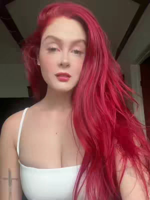 A woman with long, vibrant red hair and pale skin looks directly at the camera. She wears a white tank top. The background suggests a cozy indoor setting with visible wooden beams.