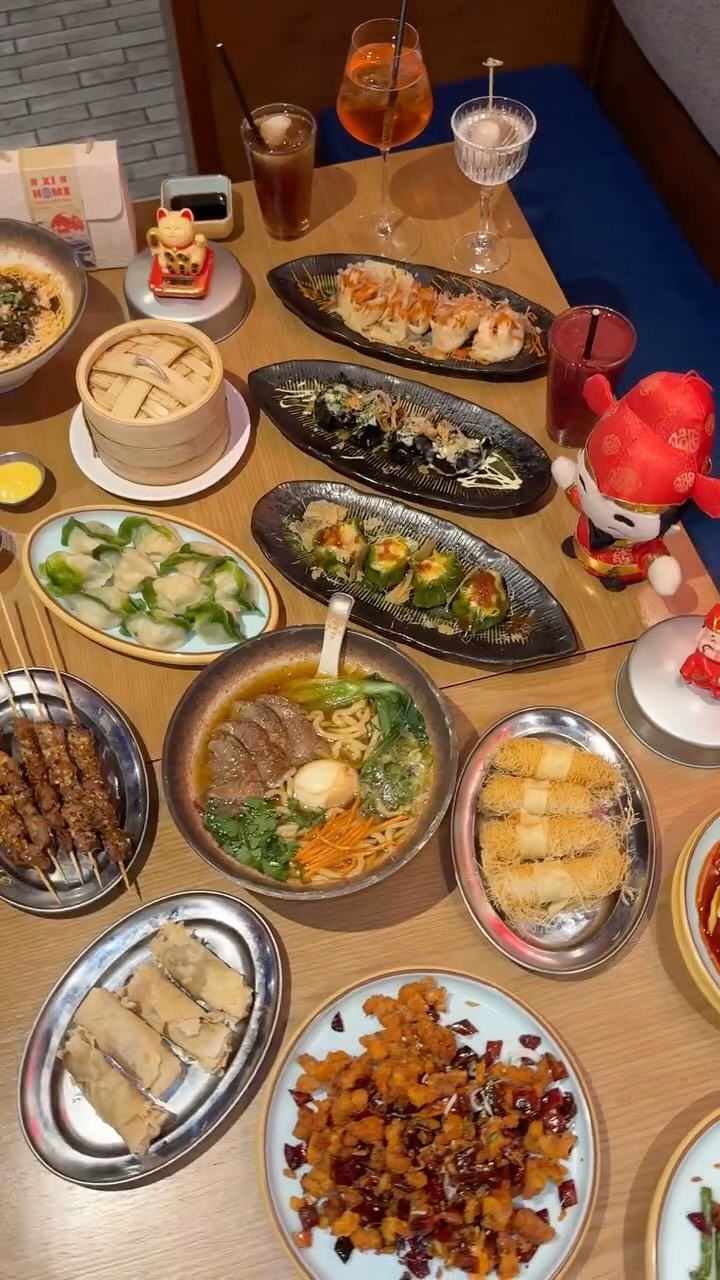 An overhead shot of a table laden with a variety of Chinese dishes. Dishes include dumplings, noodle soup, skewers, and other appetizers. Beverages and decorative items are also visible.
