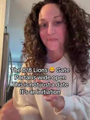 A woman with curly brown hair is the main subject, set indoors. Overlayed text in white font on a semi-transparent black box reads "The 8/8 Lions Gate Portal is wide open. This is not just a date, it's an initiation". The background is a neutral palette with a glimpse of a white kitchen. The composition is a medium shot, framed from the chest up, centered on the woman looking directly at the camera with a slight smile.