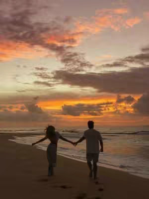 Two people, silhouetted against a colorful sunset, hold hands and walk along a beach. The sky is filled with orange, yellow, and purple hues, reflecting in the wet sand. This creates a warm and romantic scene, capturing a moment of connection and calmness.