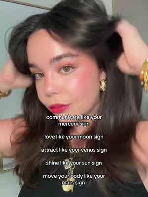 A young woman with long dark hair is shown in a close-up view, holding her hair with both hands. She wears makeup, including prominent red lipstick and blush on her cheeks, a delicate necklace, and gold earrings and bracelets. The background is out of focus, and overlaid text providing zodiac sign guidance is displayed.