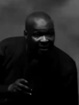 A grayscale image features a man in a dark suit, captured from the chest up with a blurred background. The man, with a shaved head, faces slightly left and appears to be speaking, possibly into a microphone. The lighting is dim, and the overall mood is serious.