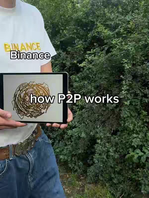 A man is holding a tablet showing a scribbled illustration in front of a background of green bushes and trees. The man is wearing a white t-shirt with text "Binance".