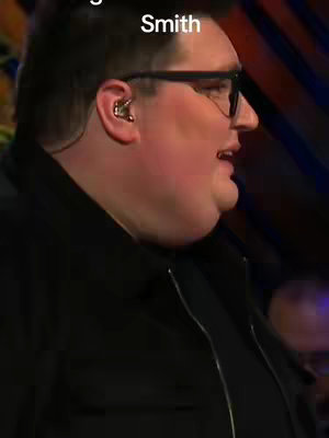 A side profile shows a man with short dark hair wearing glasses and a black jacket. He is looking to the side with his mouth slightly open, as if speaking. He has a hearing aid in his right ear.