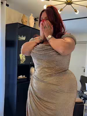 A woman in a gold, metallic dress covers her mouth with both hands in a gesture of surprise. She has red hair pulled up in a high ponytail and is standing in a brightly lit room with a mirrored wall and dark furniture.