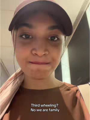 A young woman wearing a maroon cap and a brown shirt is the focus of a selfie. She has dark hair, and her expression is somewhat neutral with subtly pursed lips. The background includes blurred indoor elements and a text overlay.
