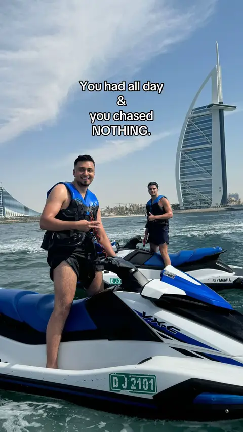 Two men are on jet skis on a body of water, possibly the ocean. In the background, a modern skyscraper with distinctive architecture is visible, suggesting a well-known city such as Dubai. The sky is clear with scattered clouds, and the overall scene projects a sense of leisure and luxury. The men are wearing life vests.