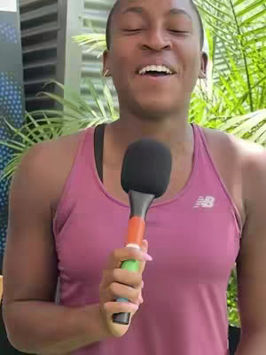 A woman with a bald head wearing a pink tank top is holding a microphone and smiling with her eyes closed. The background includes a glimpse of greenery. The lighting appears to be natural.