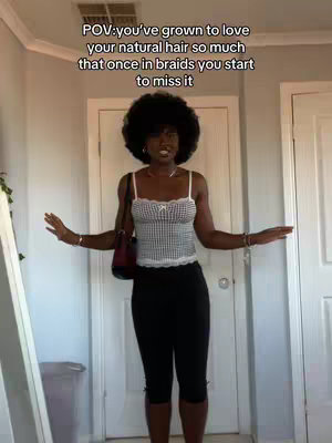 A well-lit full-body shot of a woman in a domestic setting, posing with both arms slightly outstretched. She has dark skin, an afro hairstyle, and is wearing a checkered top, black capri pants, and a small purse. Text overlay, featuring a POV-style message about natural hair. The backdrop includes a door and light-colored walls.