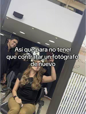 A woman is taking a selfie with a phone in her hand. She is in what looks like a salon. There is a man in the background smiling. Text has been added to the image in a white font.