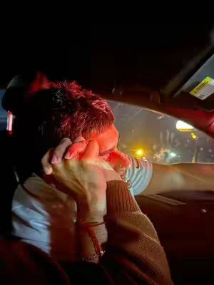 A dimly lit shot inside a car at night, focusing on two people holding hands. The hands of the two individuals are the main focus, with one arm draped over the other. The lighting suggests the scene is lit by the car's interior lights and external sources, creating a low-contrast environment. The shot is framed to emphasize intimacy and connection.
