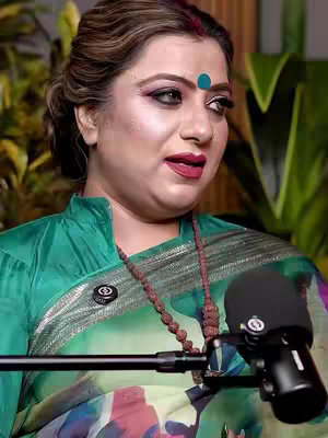 A woman with a painted blue dot on her forehead is speaking, with a microphone in front of her. She is wearing a green and patterned sari and a necklace of brown beads. A microphone is positioned in front of her, and the background shows leafy green plants.
